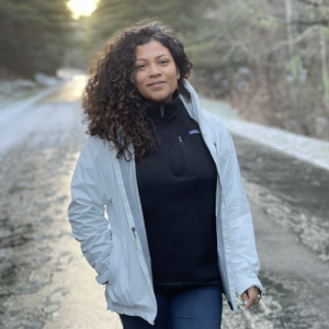 Iris is pictured standing in front of a snowy path and wearing a light grey jacket over a black jacket. Her curly brown hair is swept to her right side.