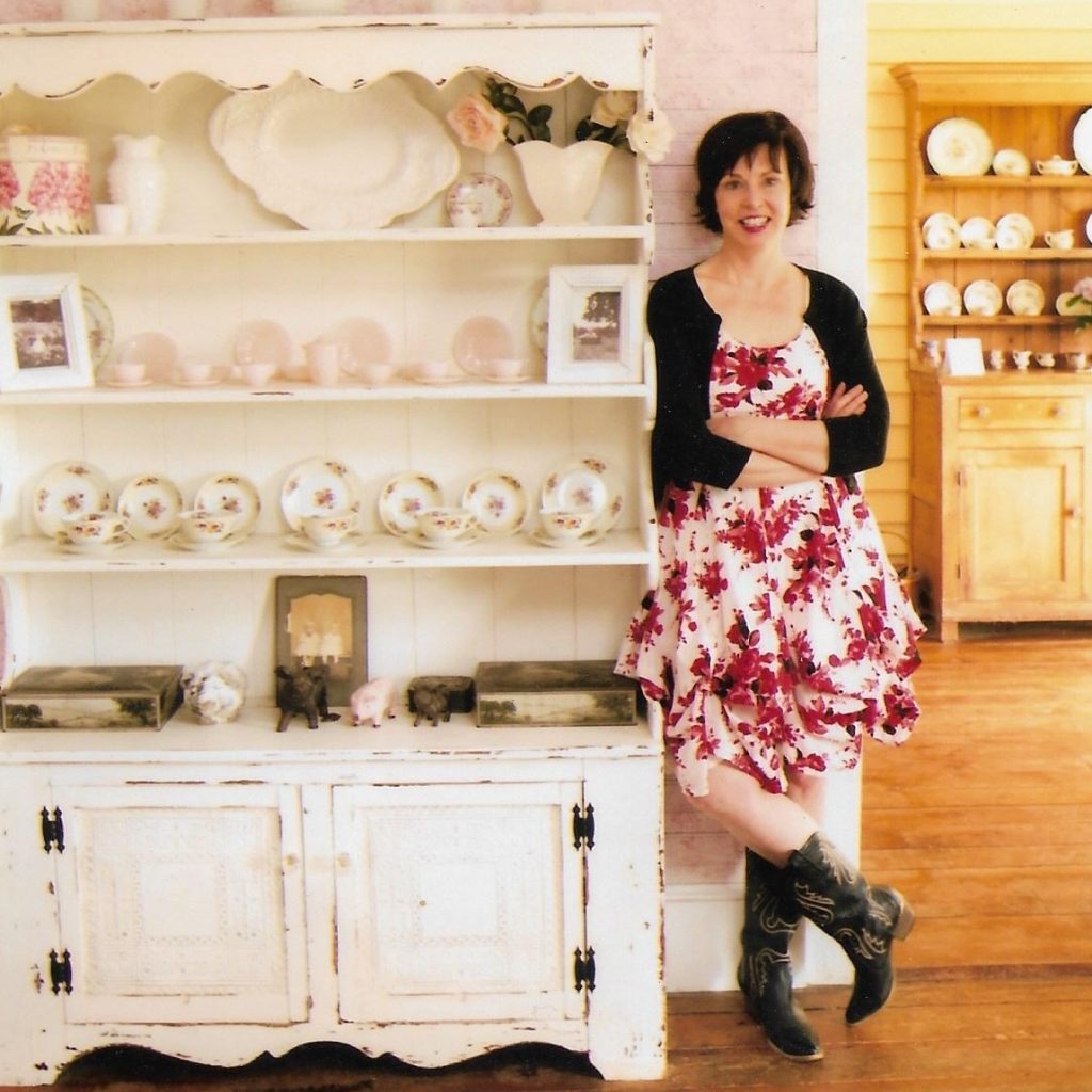 Woman wearing a red flowered dress, a black cardigan and cowboy boots standing next to a hutch.