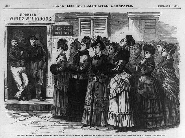 A crowd of women praying outside of a saloon while men smoke and laugh in the doorway.