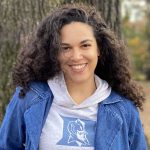 headshot of Amber Richardson, Senior Program Coordinator for the Center for Pathway Programs. Amber is pictured smiling in front of a tree, wearing a Duke sweatshirt under a denim jacket. She has long dark curly hair.