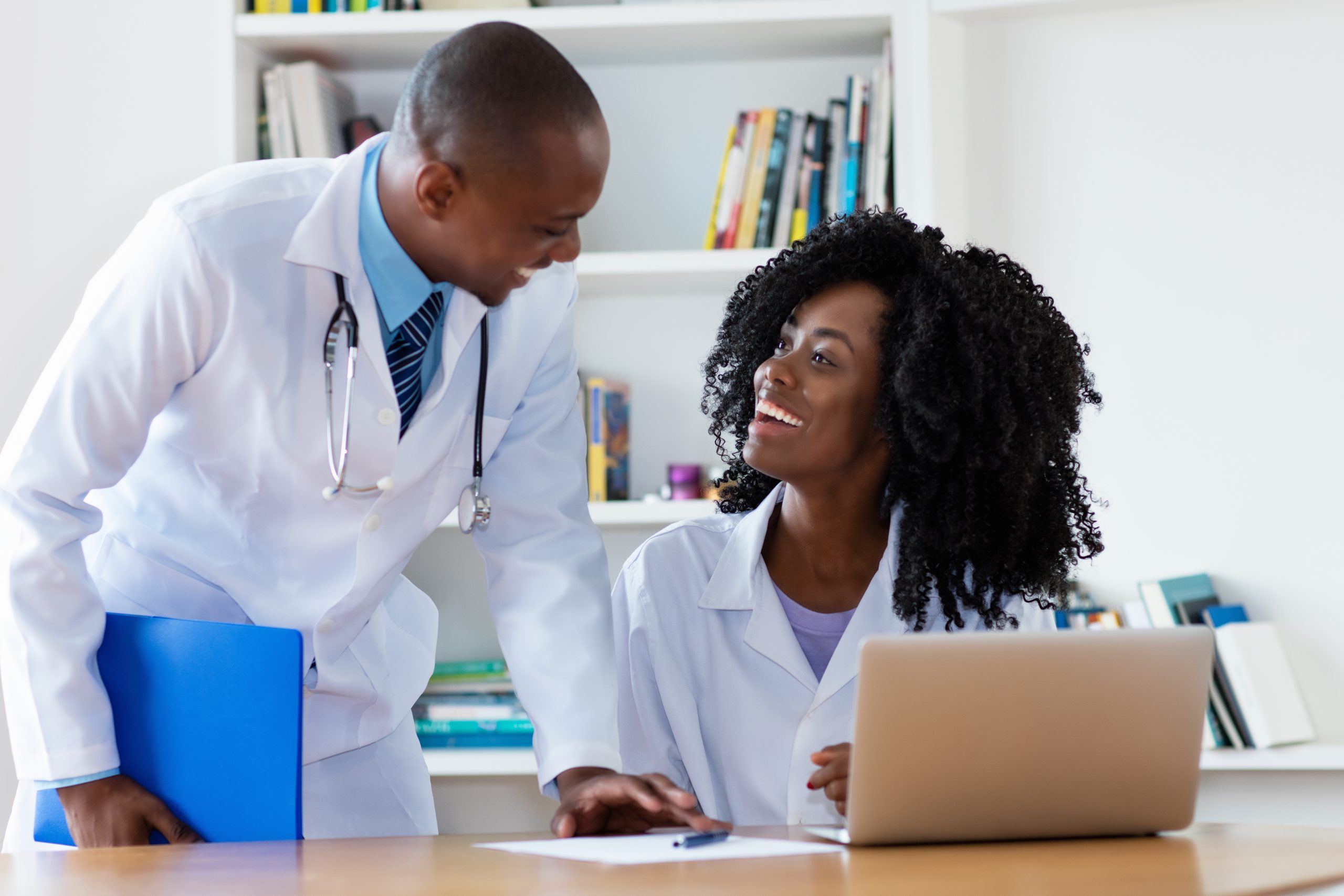 A Black male chief physician leans over a table and smiles at a smiling Black woman doctor as she is seated behind the same table with a laptop in front of her.