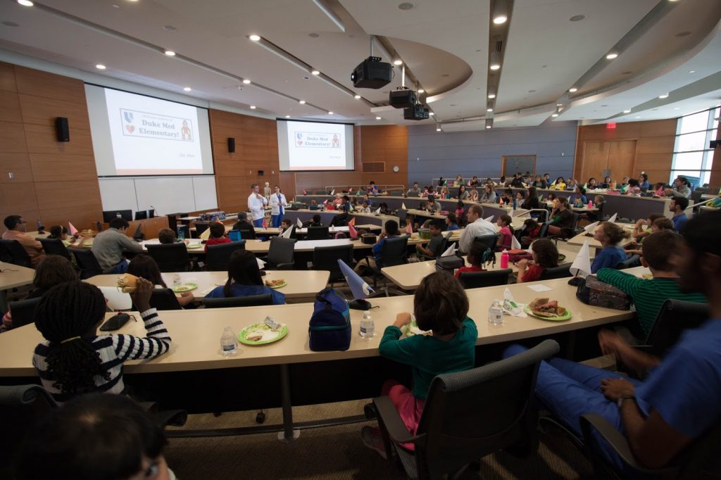 A lecturer addresses a lecture hall full of elementary students who look on and eat lunch as they listen to the presentation.