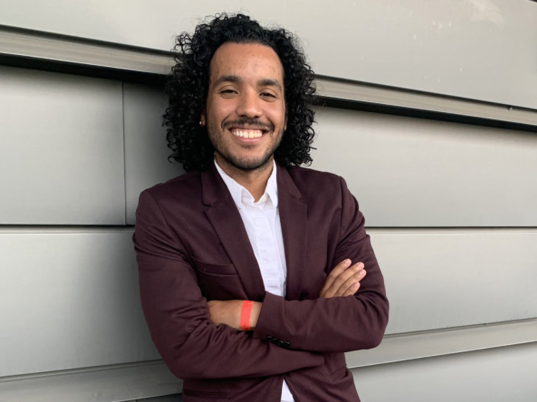 Daniel Colón Hidalgo, MD MPH, is pictured smiling and standing in front of a grey wall. Daniel has curly dark brown shoulder length hair and is smiling with arms crossed. He wears a deep red blazer.