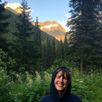 Woman smiles with eyes closed and hood on, with a mountain and forest in the background 