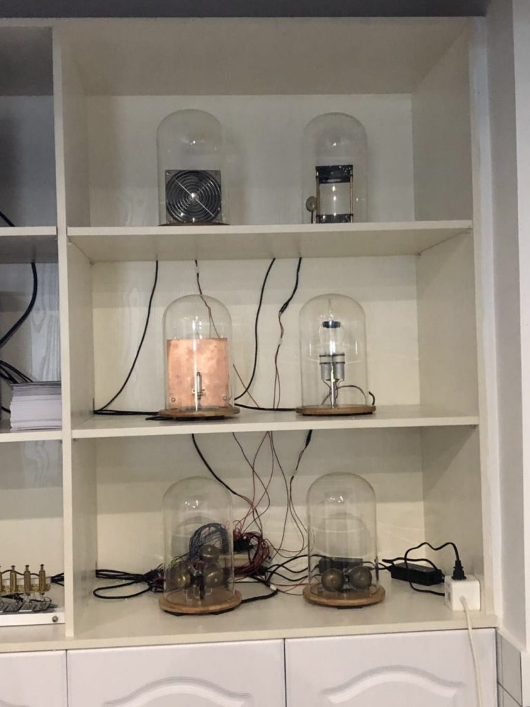 Bell jars with bells or other percussive instruments in them rest on a shelf. Wires connect them to a power source and something offscreen.