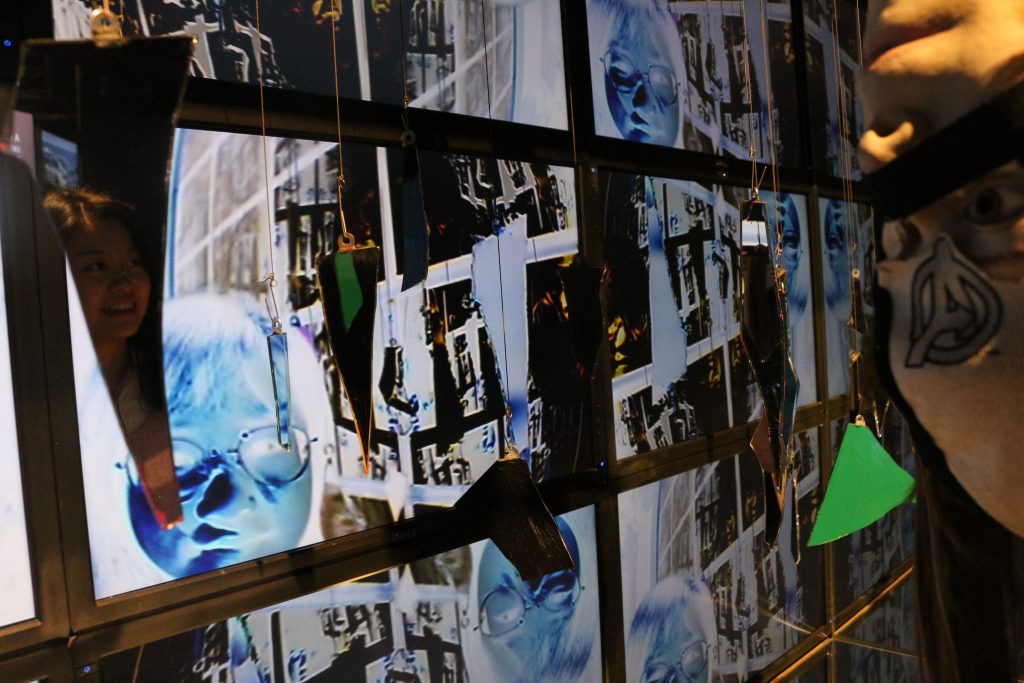 Mirror shards hang in front of screens displaying live video of the viewers, with the colors inverted. On the right, a mannequin head hangs upside-down. The Avengers logo is visible on a piece of cloth on the head.