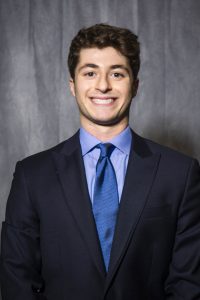 indoor portrait of Alex wearing a navy suit, blue shirt, blue tie, with grey background