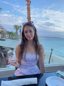 outdoor shot of Alice at a restaurant on the beach wearing a white dress and holding a drink