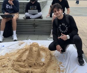 outdoor photo of Ardil kneeling down and pointing at a sand castle wearing all black