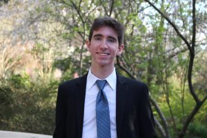 outdoor head shot of Chris wearing a black suit and blue tie with trees in the background