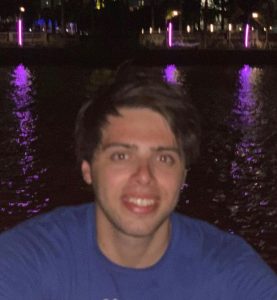 casual shot of Thomas wearing a blue shirt at night in front of a river with purple lights in the background