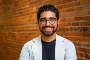 indoor head shot of Wanghley wearing white blazer and black shirt with brick wall background