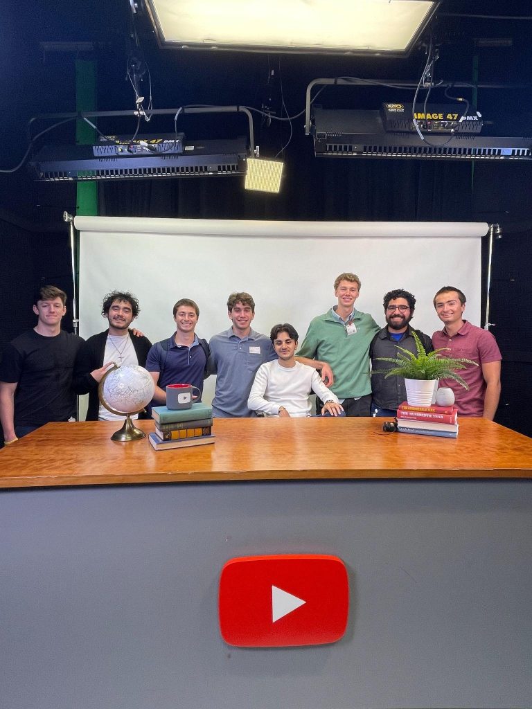 Eight students on a soundstage behind a desk with the YouTube logo and props