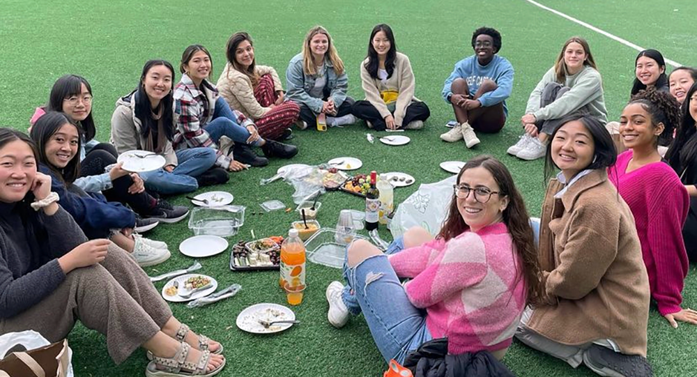 Group of young women sitting in a circle outdoors and eating food and drink with paper plates and cups.