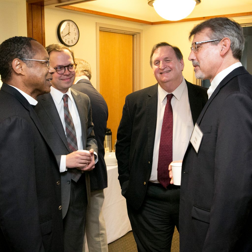 There are four men talking in a circle. From left to right, a Black man, a younger White man with sporty glasses, a older White man, and another White man.