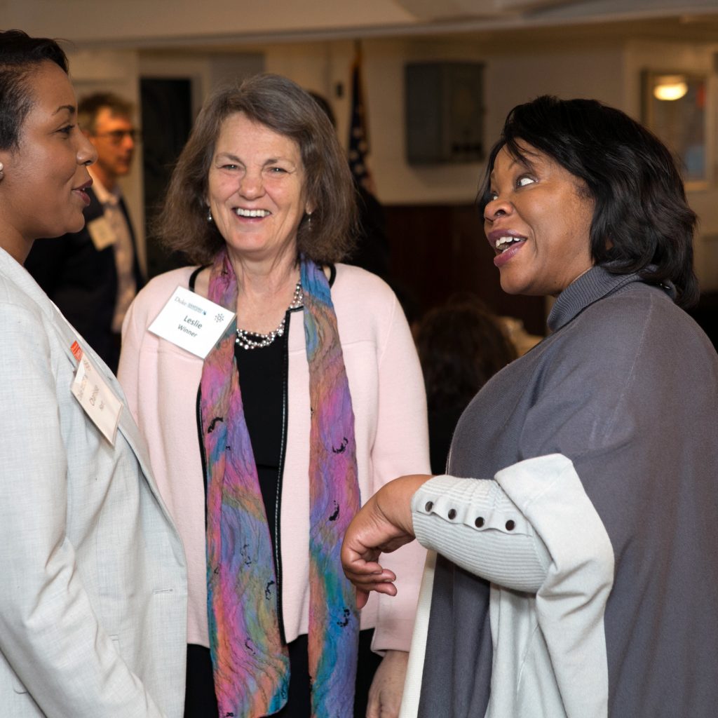 Three women speak. From the left to the right, there is a black woman with a tan blazer, there is a white woman with a multicolor scarf and pink cardigan, and a Black woman wearing a grey poncho.