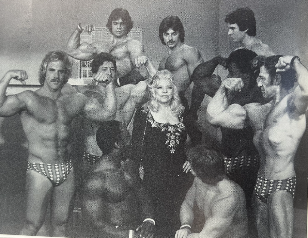 A woman stands, hand on her hip, surrounded by men in speedos flexing their arms.